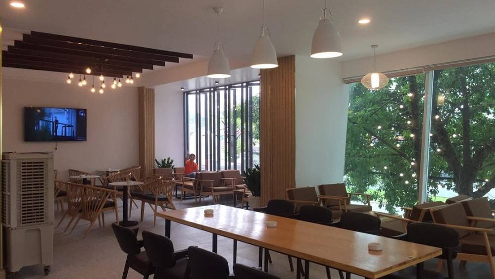 Qu&aacute;n c&agrave; ph&ecirc; hiện đại với b&agrave;n ghế gỗ, đ&egrave;n treo trần, cửa sổ lớn nh&igrave;n ra c&acirc;y xanh b&ecirc;n ngo&agrave;i.