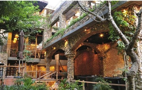 Quán cà phê với kiến trúc đá và gạch, không gian mở, cây xanh xung quanh, ánh sáng vàng ấm áp.