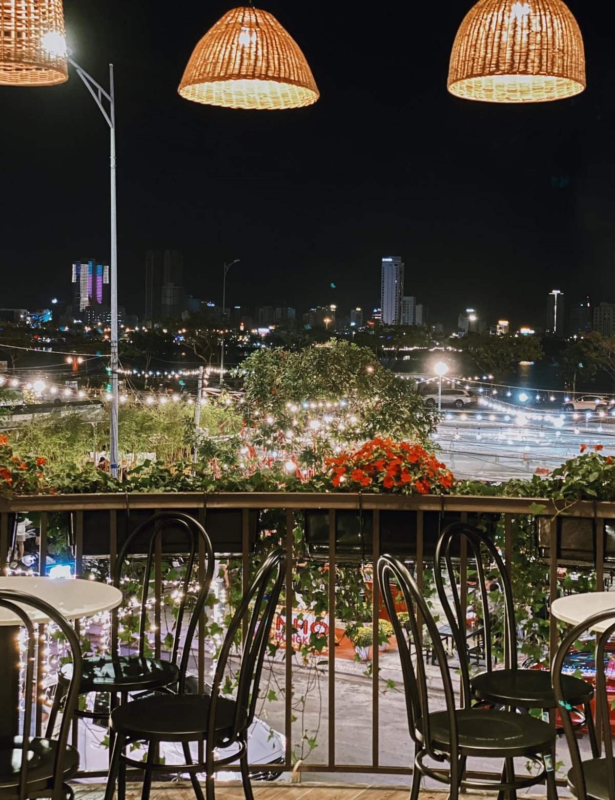 Qu&aacute;n c&agrave; ph&ecirc; ban đ&ecirc;m với đ&egrave;n treo, b&agrave;n ghế ngo&agrave;i trời, c&acirc;y xanh v&agrave; th&agrave;nh phố s&aacute;ng đ&egrave;n ph&iacute;a xa.