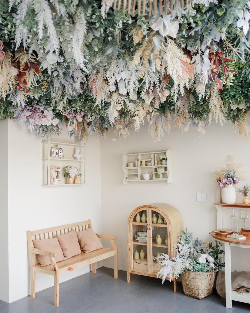 G&oacute;c ph&ograve;ng trang tr&iacute; với hoa treo tr&ecirc;n trần, ghế gỗ, tủ k&iacute;nh v&agrave; kệ trưng b&agrave;y lọ hoa v&agrave; vật dụng trang tr&iacute;.