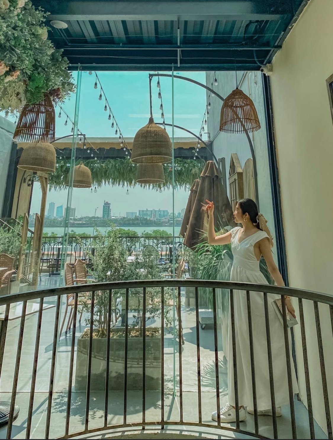 Người phụ nữ đứng tr&ecirc;n ban c&ocirc;ng với cảnh quan th&agrave;nh phố v&agrave; hồ nước, trang tr&iacute; bằng đ&egrave;n v&agrave; c&acirc;y xanh.