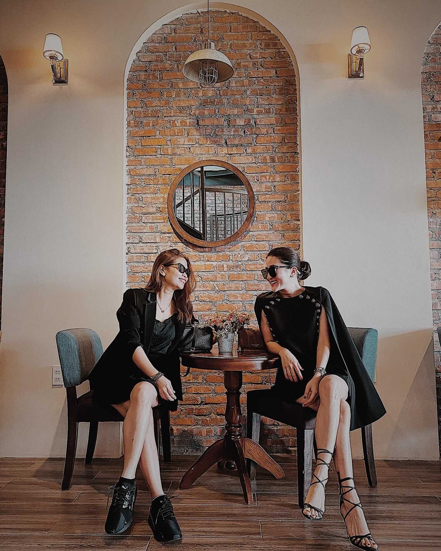 Hai người ngồi tr&ograve; chuyện tại qu&aacute;n c&agrave; ph&ecirc;, mặc trang phục đen, ph&iacute;a sau l&agrave; tường gạch v&agrave; gương tr&ograve;n.