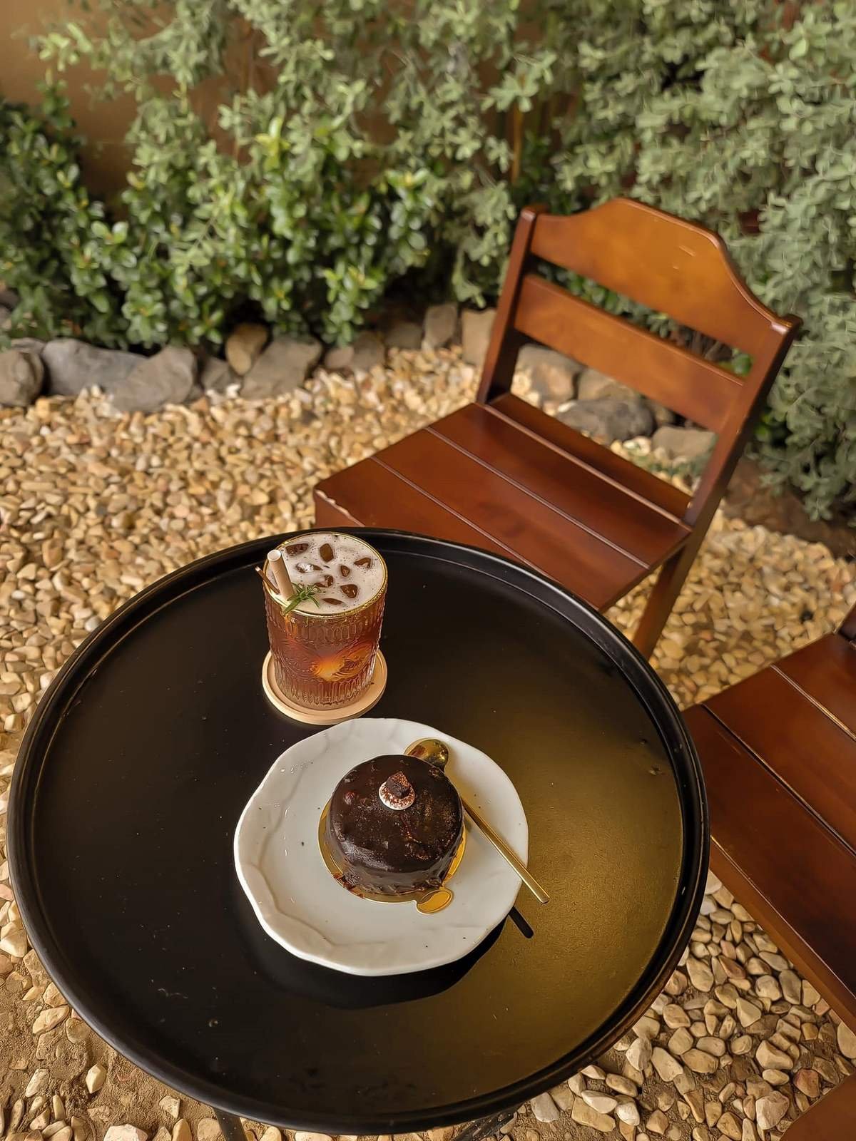 Ly đồ uống v&agrave; b&aacute;nh ngọt tr&ecirc;n b&agrave;n tr&ograve;n đen, xung quanh l&agrave; ghế gỗ v&agrave; nền đ&aacute; cuội, c&oacute; c&acirc;y xanh ph&iacute;a sau.