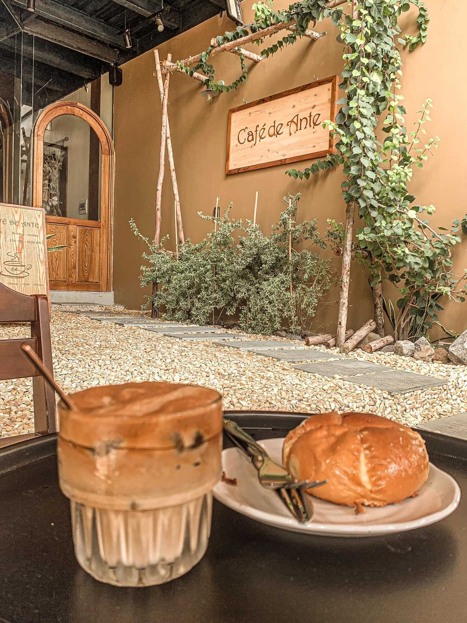 Ly c&agrave; ph&ecirc; v&agrave; b&aacute;nh tr&ecirc;n b&agrave;n, nền l&agrave; qu&aacute;n c&agrave; ph&ecirc; với bảng hiệu v&agrave; c&acirc;y xanh trang tr&iacute;.