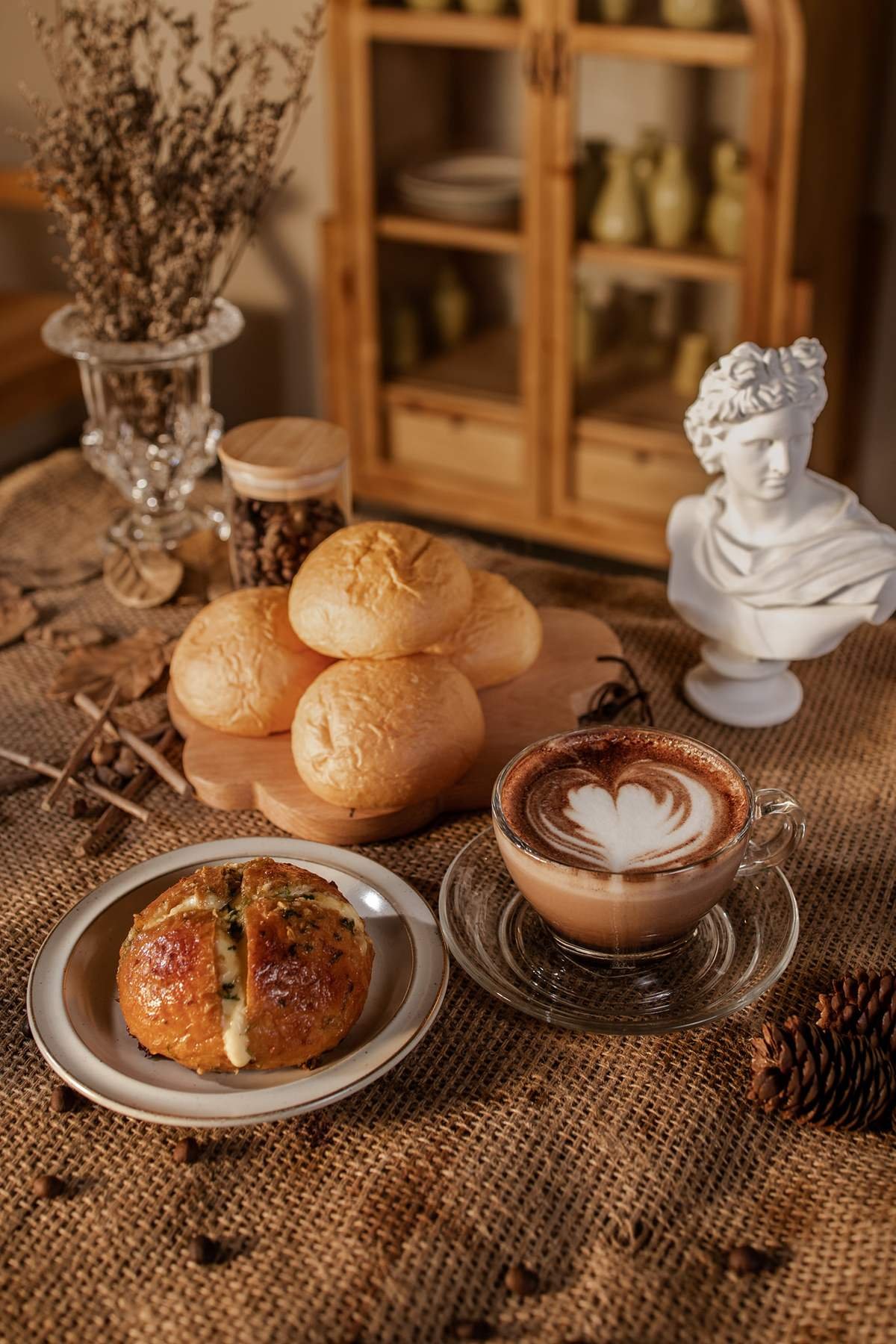 B&agrave;n ăn với b&aacute;nh m&igrave;, c&agrave; ph&ecirc; latte, tượng b&aacute;n th&acirc;n v&agrave; lọ hoa kh&ocirc;, tạo kh&ocirc;ng gian ấm c&uacute;ng, cổ điển.