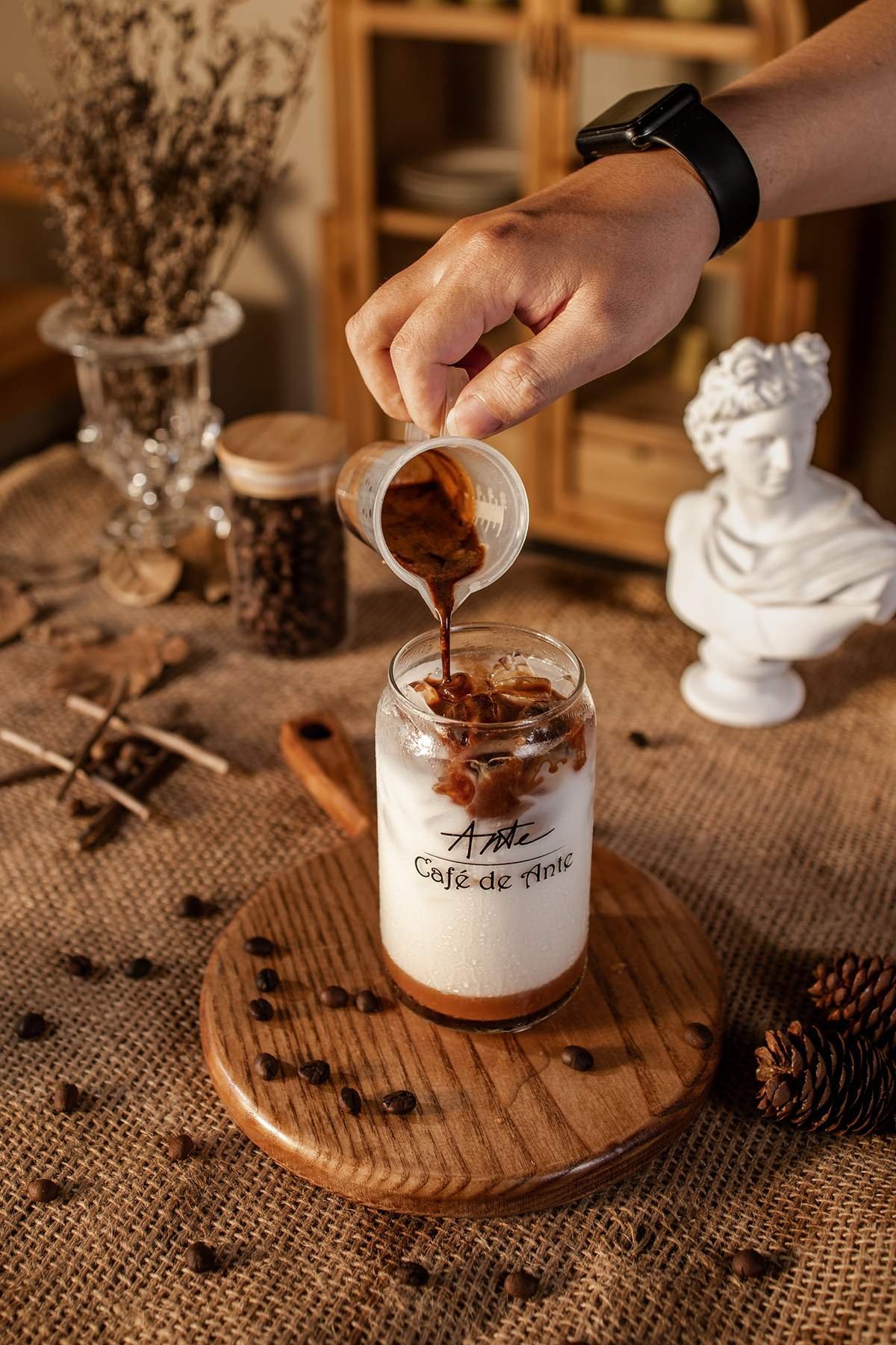 Tay đang r&oacute;t c&agrave; ph&ecirc; từ ly nhựa v&agrave;o ly thủy tinh với đ&aacute;, tr&ecirc;n b&agrave;n c&oacute; hạt c&agrave; ph&ecirc; v&agrave; đồ trang tr&iacute;.