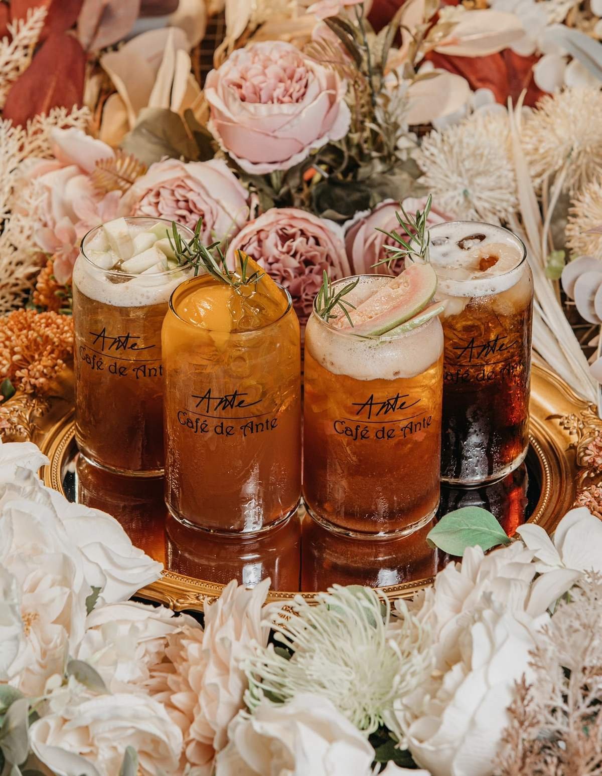 Bốn ly đồ uống tr&ecirc;n khay v&agrave;ng, xung quanh l&agrave; hoa hồng v&agrave; hoa trang tr&iacute;.