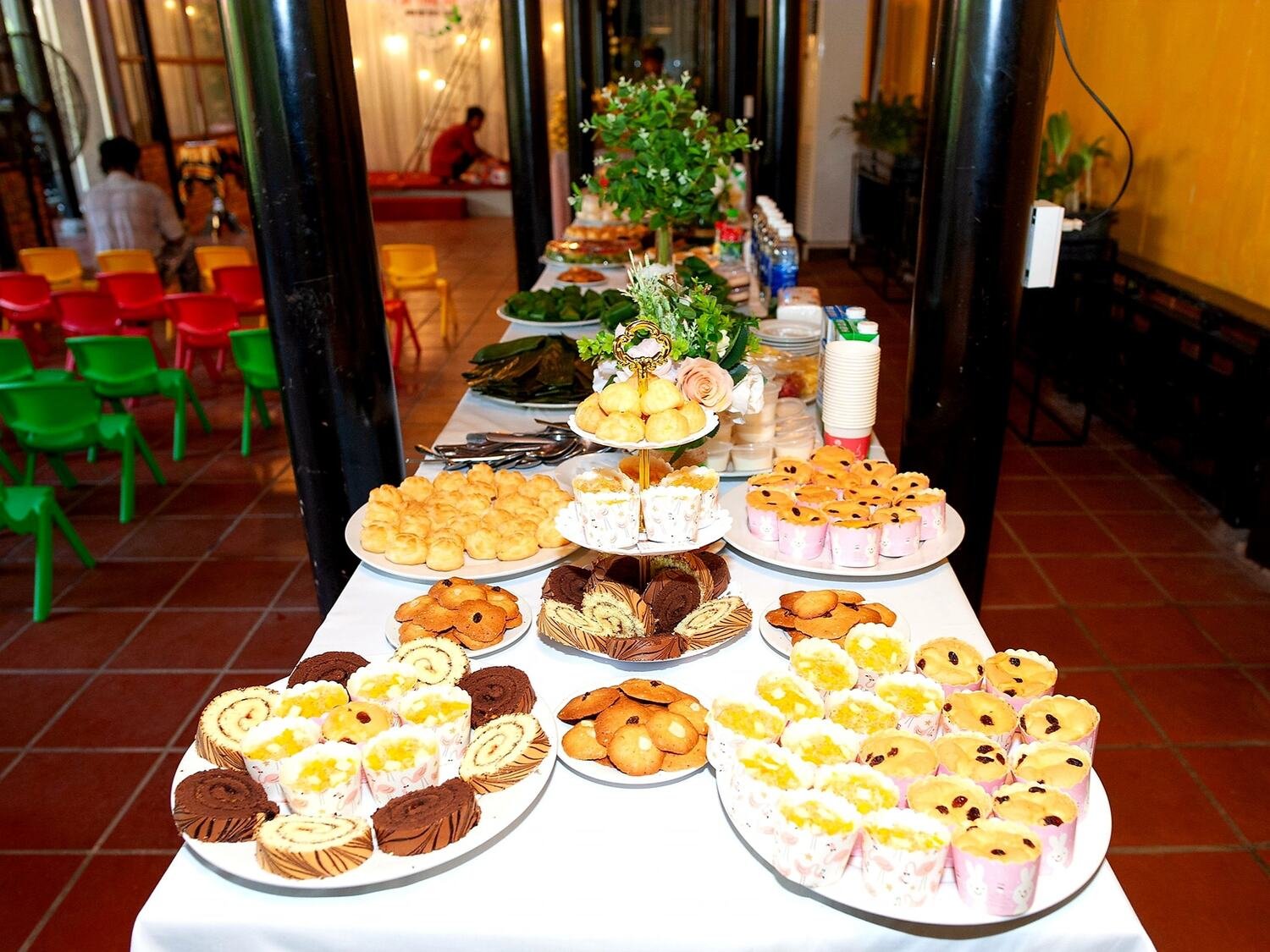 B&agrave;n tiệc với nhiều loại b&aacute;nh ngọt v&agrave; tr&aacute;i c&acirc;y, ph&iacute;a sau l&agrave; ghế nhựa m&agrave;u sắc v&agrave; kh&ocirc;ng gian trang tr&iacute; ấm c&uacute;ng.