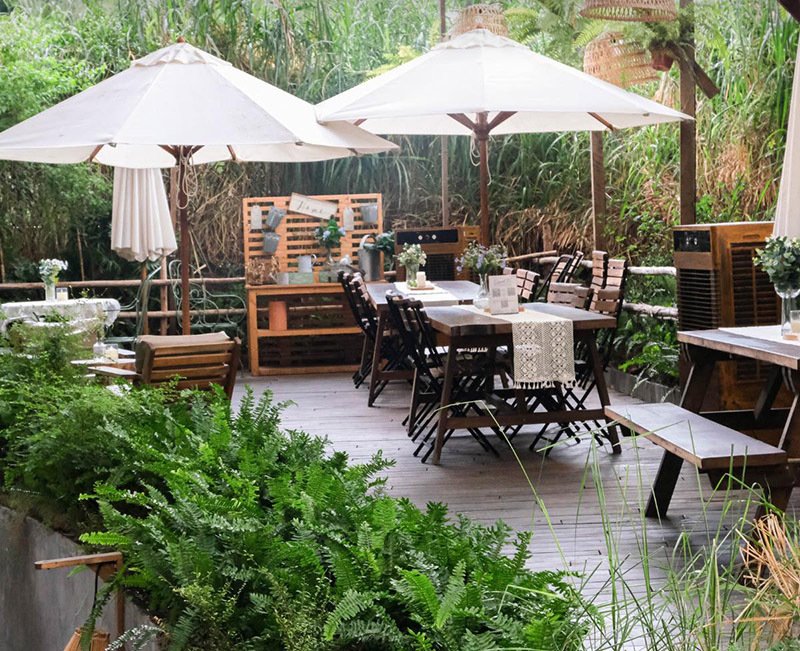 Khu vực ăn uống ngo&agrave;i trời với b&agrave;n ghế gỗ, &ocirc; che nắng trắng, c&acirc;y xanh xung quanh tạo kh&ocirc;ng gian thoải m&aacute;i.
