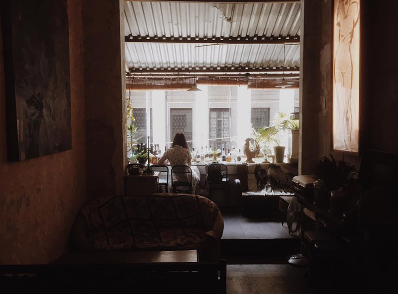 Căn ph&ograve;ng với &aacute;nh s&aacute;ng tự nhi&ecirc;n, một người ngồi b&ecirc;n cửa sổ lớn, xung quanh l&agrave; nhiều đồ vật v&agrave; c&acirc;y cảnh.