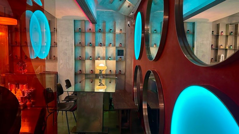 Qu&aacute;n c&agrave; ph&ecirc; với thiết kế hiện đại, &aacute;nh s&aacute;ng xanh v&agrave; đỏ, b&agrave;n ghế tối giản, kệ trưng b&agrave;y nhiều chai lọ.