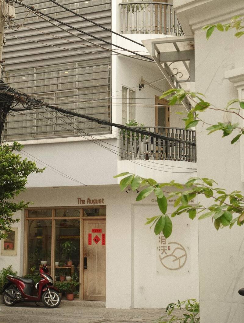 Mặt tiền qu&aacute;n c&agrave; ph&ecirc; với cửa k&iacute;nh, c&acirc;y xanh v&agrave; xe m&aacute;y đậu ph&iacute;a trước, d&acirc;y điện chạy ngang qua ph&iacute;a tr&ecirc;n.