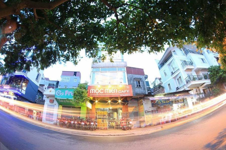 cafe-sach-ha-noi-8 Một quán cà phê với bảng hiệu sáng đèn nằm trên con phố sầm uất, xung quanh là các cửa hàng khác và cây xanh.