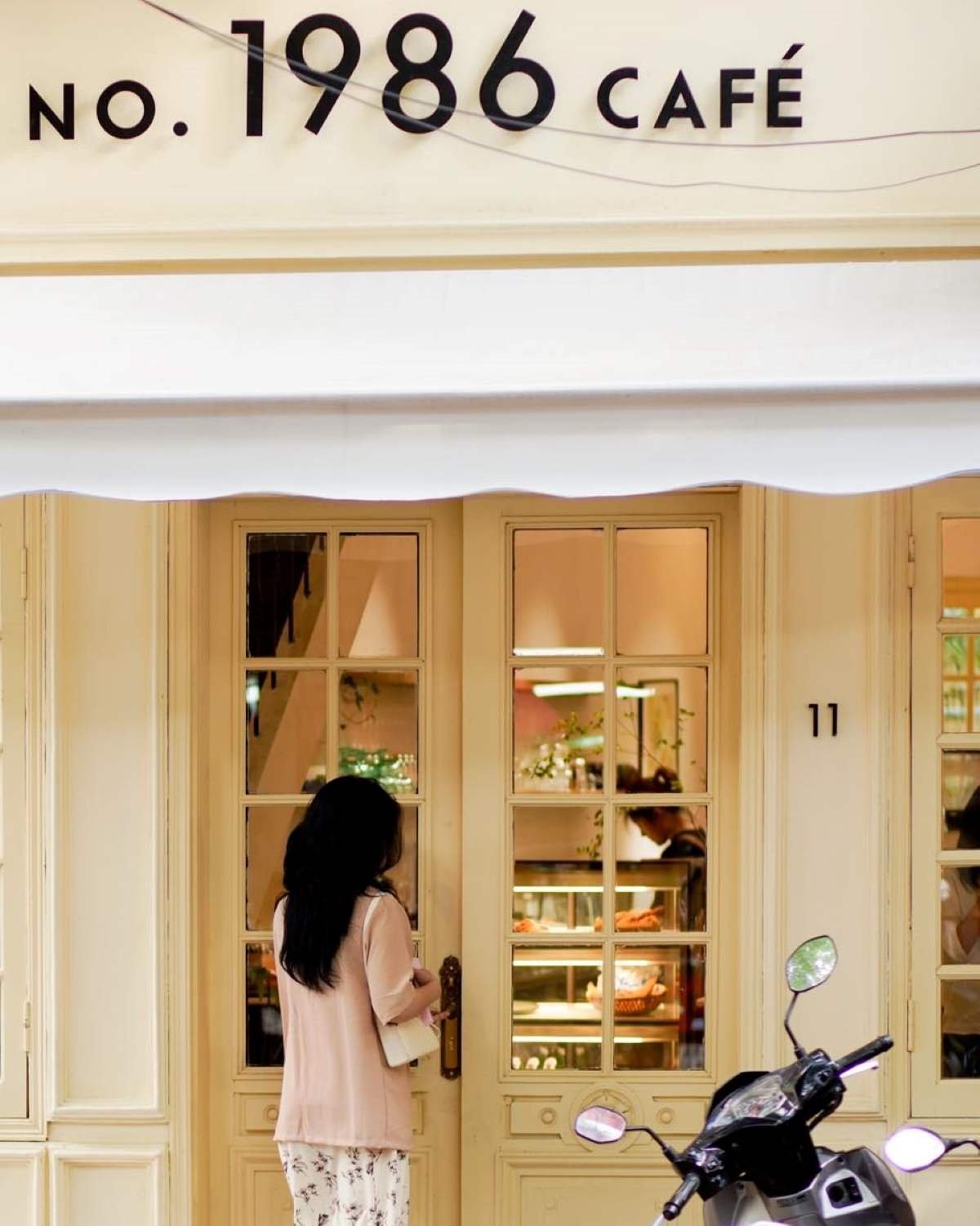 Người đứng trước cửa qu&aacute;n c&agrave; ph&ecirc; NO. 1986, cạnh xe m&aacute;y, b&ecirc;n trong c&oacute; kệ trưng b&agrave;y b&aacute;nh ngọt.