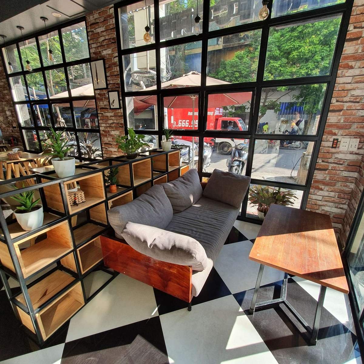 Qu&aacute;n c&agrave; ph&ecirc; với ghế sofa x&aacute;m, b&agrave;n gỗ, kệ trang tr&iacute; v&agrave; cửa sổ lớn nh&igrave;n ra đường phố s&ocirc;i động.