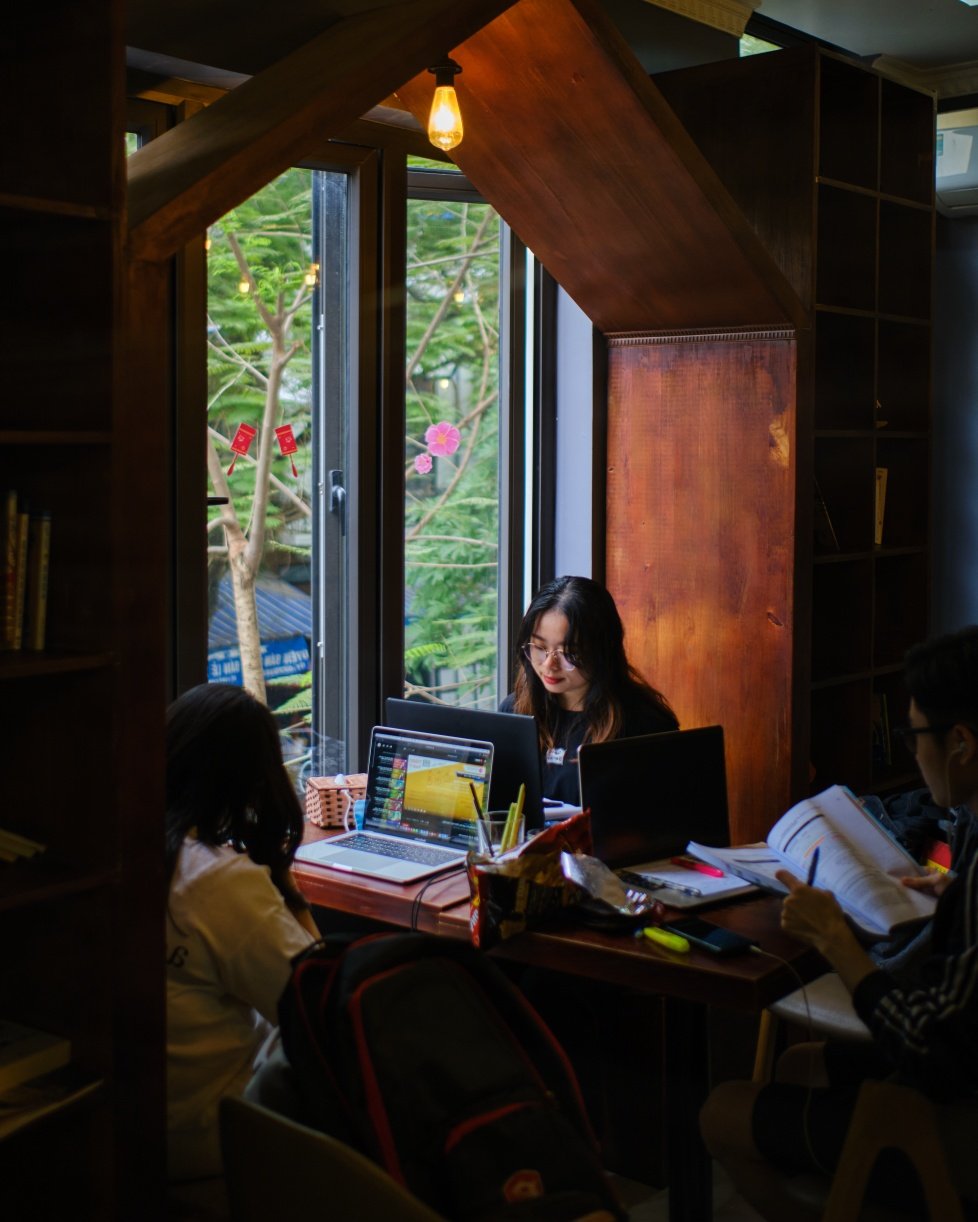 Ba người ngồi học tại một g&oacute;c cửa sổ, sử dụng m&aacute;y t&iacute;nh x&aacute;ch tay v&agrave; đọc s&aacute;ch, xung quanh c&oacute; nhiều c&acirc;y xanh.