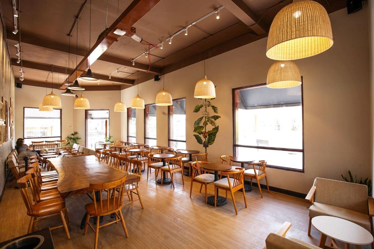 Qu&aacute;n c&agrave; ph&ecirc; với b&agrave;n ghế gỗ, đ&egrave;n treo &aacute;nh s&aacute;ng v&agrave;ng, cửa sổ lớn v&agrave; c&acirc;y xanh trang tr&iacute; tr&ecirc;n tường.