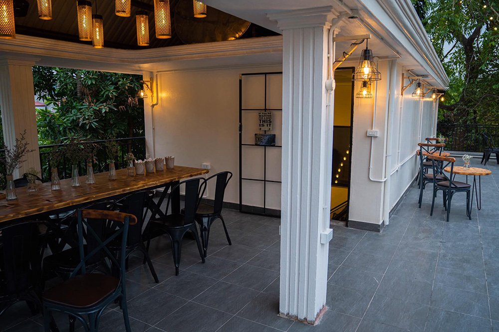 Khu vực ngo&agrave;i trời của qu&aacute;n c&agrave; ph&ecirc; với b&agrave;n ghế gỗ, đ&egrave;n trang tr&iacute; v&agrave; c&acirc;y xanh xung quanh.