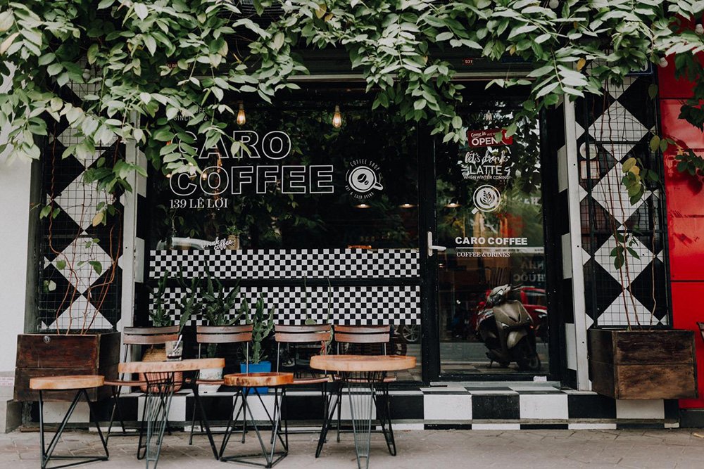 Mặt tiền qu&aacute;n c&agrave; ph&ecirc; với c&acirc;y xanh phủ k&iacute;n, b&agrave;n ghế ngo&agrave;i trời v&agrave; cửa k&iacute;nh c&oacute; logo qu&aacute;n.