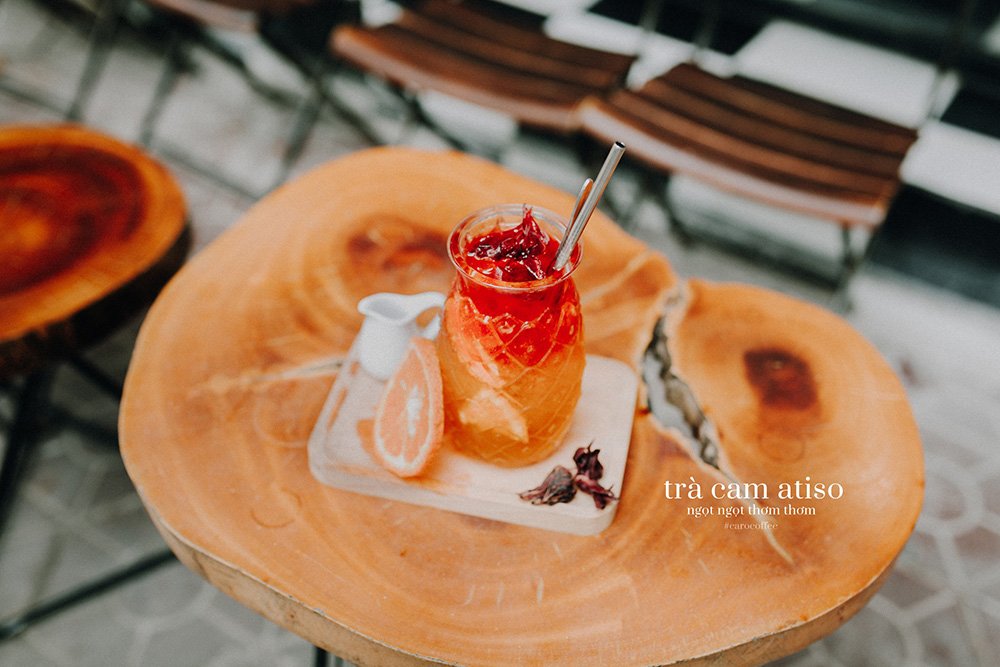 Ly tr&agrave; cam atiso tr&ecirc;n b&agrave;n gỗ, k&egrave;m l&aacute;t cam v&agrave; b&igrave;nh nhỏ, trang tr&iacute; hoa kh&ocirc;.