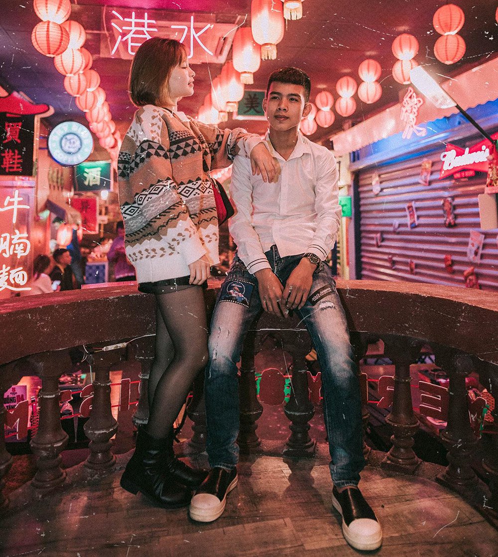 Hai người đứng tr&ecirc;n cầu, xung quanh l&agrave; đ&egrave;n lồng đỏ v&agrave; biển hiệu neon trong một khu phố.