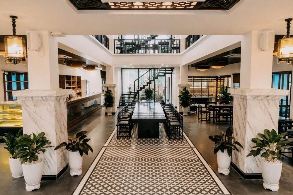 Kh&ocirc;ng gian nội thất sang trọng với b&agrave;n ghế gỗ, s&agrave;n l&oacute;t gạch hoa văn v&agrave; c&acirc;y cảnh trang tr&iacute;, c&oacute; cầu thang ở giữa.
