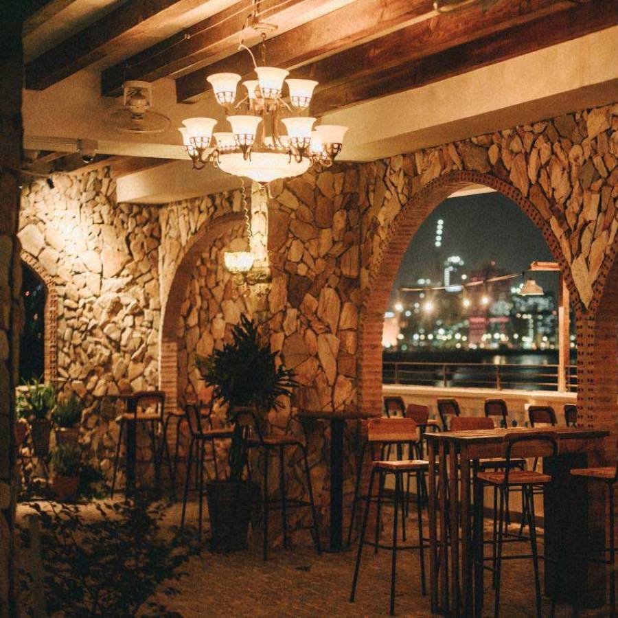 Qu&aacute;n c&agrave; ph&ecirc; với tường đ&aacute; v&agrave; đ&egrave;n ch&ugrave;m, b&agrave;n ghế gỗ, nh&igrave;n ra khung cảnh th&agrave;nh phố về đ&ecirc;m.