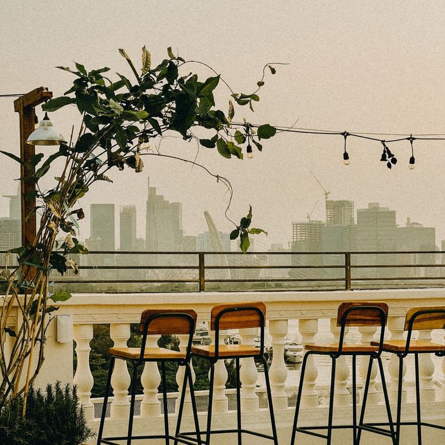 Ghế cao v&agrave; b&agrave;n tr&ecirc;n ban c&ocirc;ng với c&acirc;y xanh, nh&igrave;n ra khung cảnh th&agrave;nh phố v&agrave; bầu trời u &aacute;m.