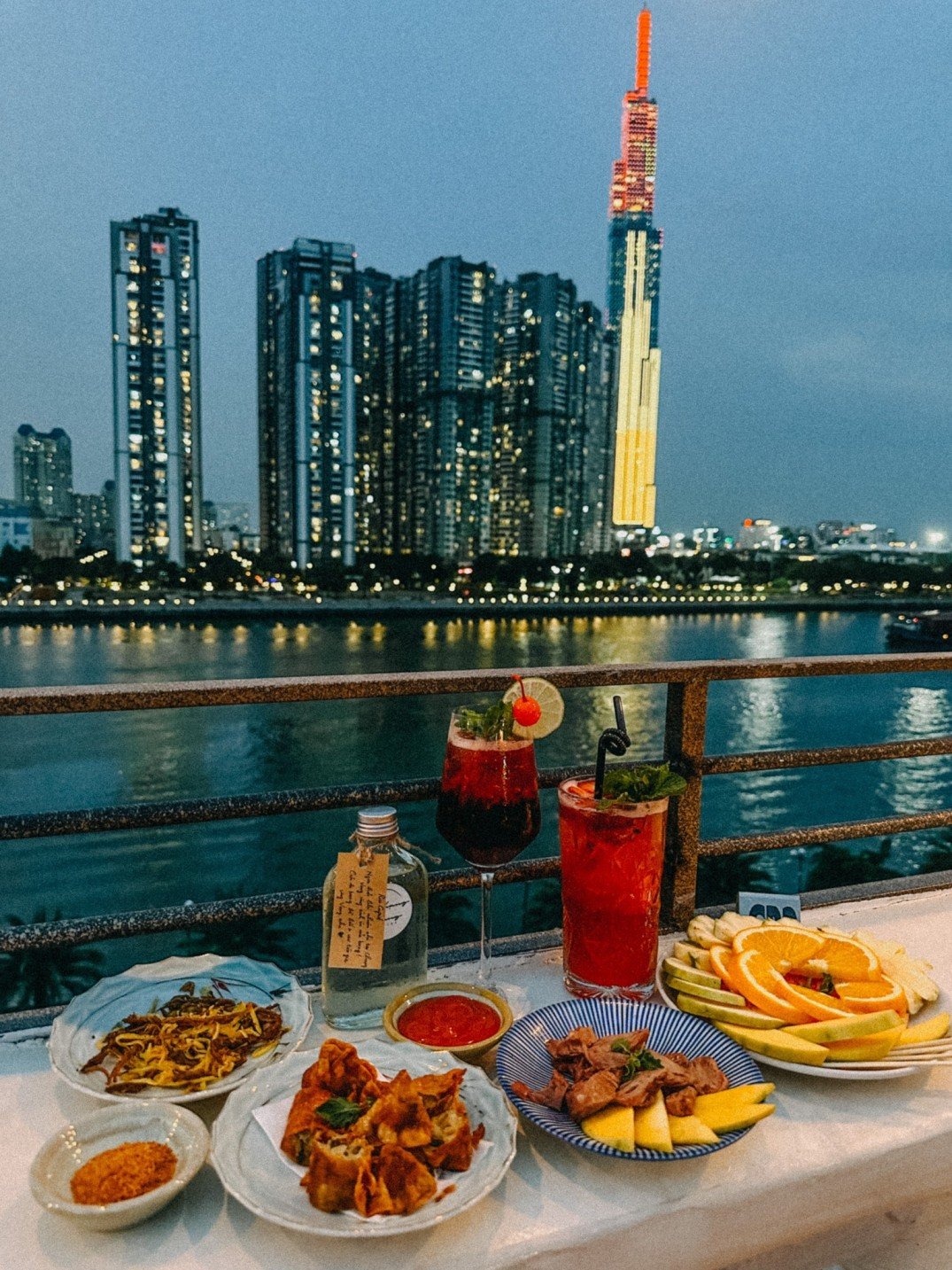 B&agrave;n ăn ngo&agrave;i trời với nhiều m&oacute;n ăn v&agrave; đồ uống, ph&iacute;a sau l&agrave; cảnh th&agrave;nh phố v&agrave; t&ograve;a nh&agrave; cao b&ecirc;n s&ocirc;ng v&agrave;o buổi tối.