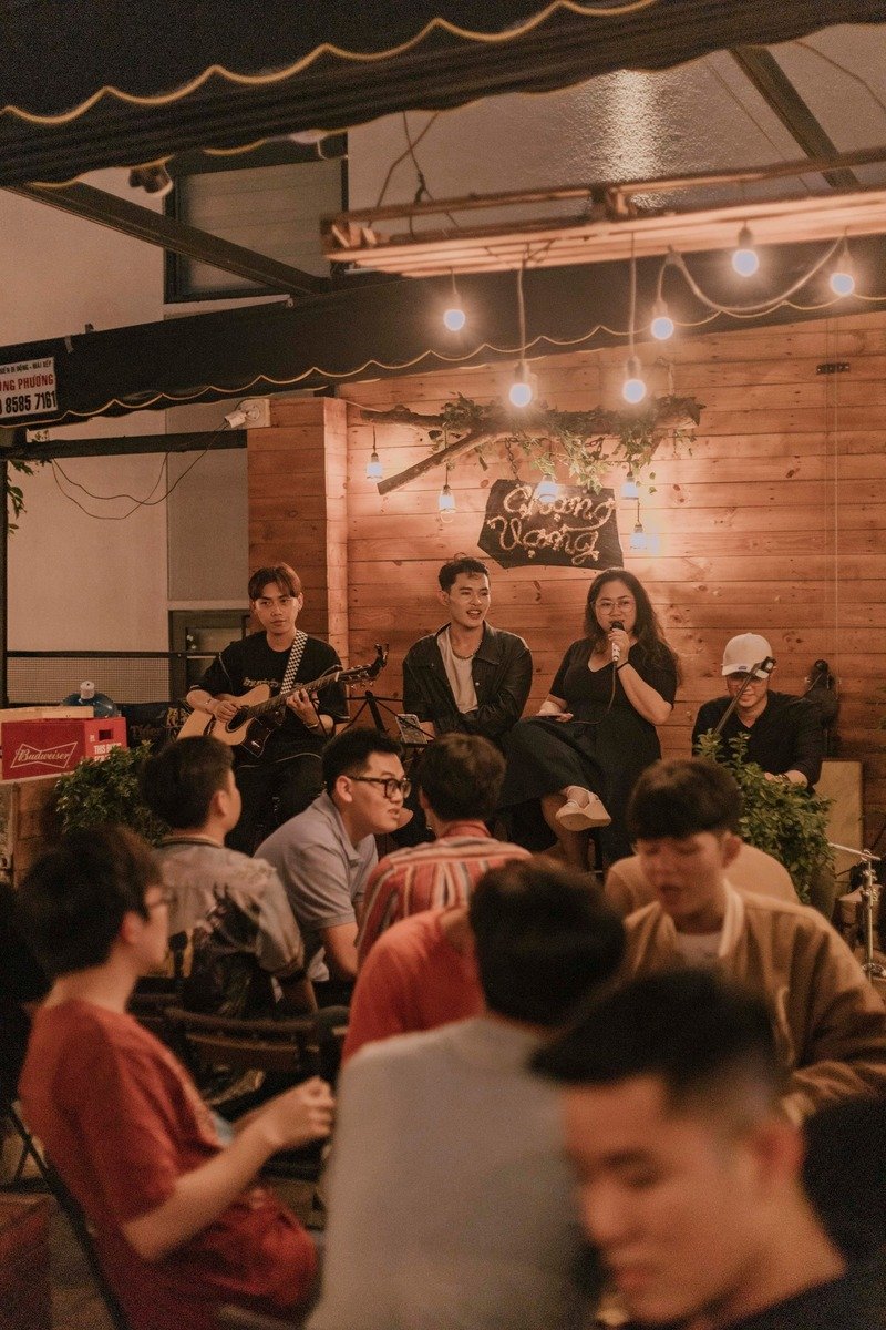 Ban nhạc biểu diễn trong qu&aacute;n c&agrave; ph&ecirc; với kh&aacute;n giả ngồi xung quanh, &aacute;nh s&aacute;ng ấm &aacute;p v&agrave; trang tr&iacute; gỗ.