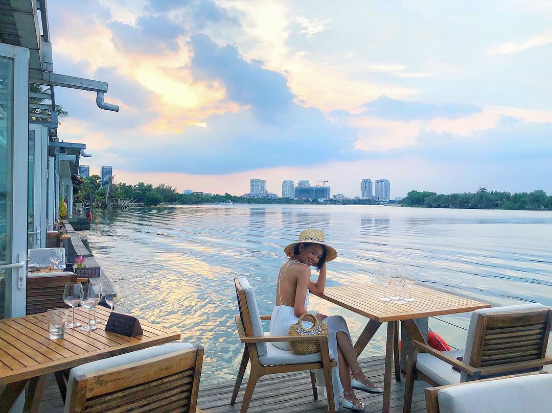 Người ngồi bên bàn cạnh bờ sông, khung cảnh hoàng hôn, tòa nhà cao tầng phía xa, không gian thư giãn và yên bình.