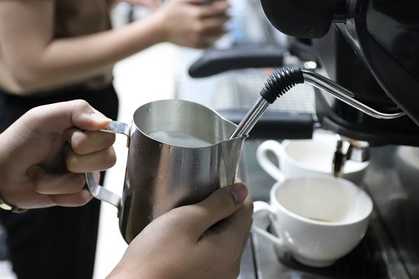 Người pha chế đang đánh sữa bằng vòi hơi nước từ máy pha cà phê, với hai tách trên quầy.