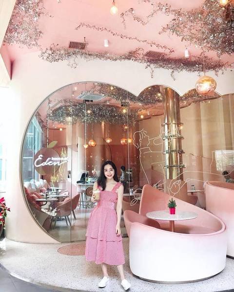 Người phụ nữ đứng trong qu&aacute;n c&agrave; ph&ecirc; trang tr&iacute; t&ocirc;ng hồng, c&oacute; ghế sofa v&agrave; b&agrave;n tr&ograve;n, trần nh&agrave; treo đ&egrave;n lấp l&aacute;nh.