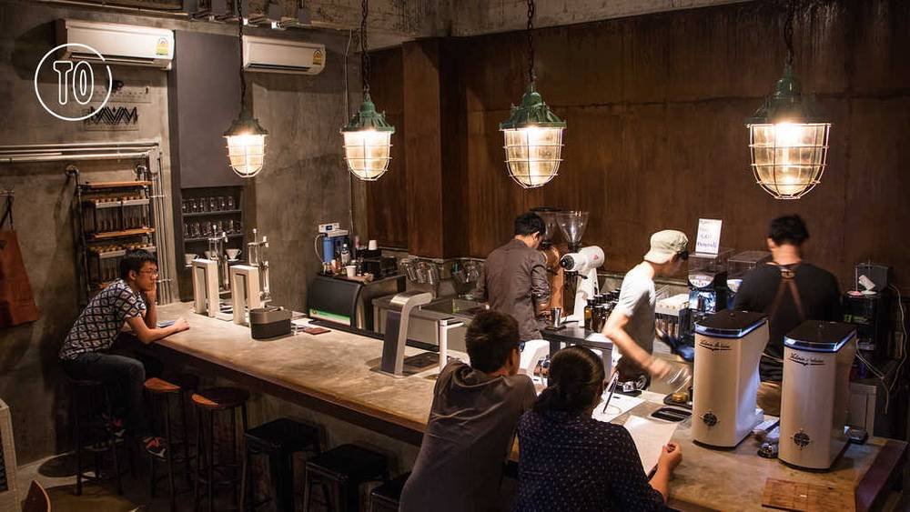 Qu&aacute;n c&agrave; ph&ecirc; với &aacute;nh s&aacute;ng ấm &aacute;p, mọi người ngồi v&agrave; đứng quanh quầy pha chế, b&agrave;n ghế gỗ đơn giản.