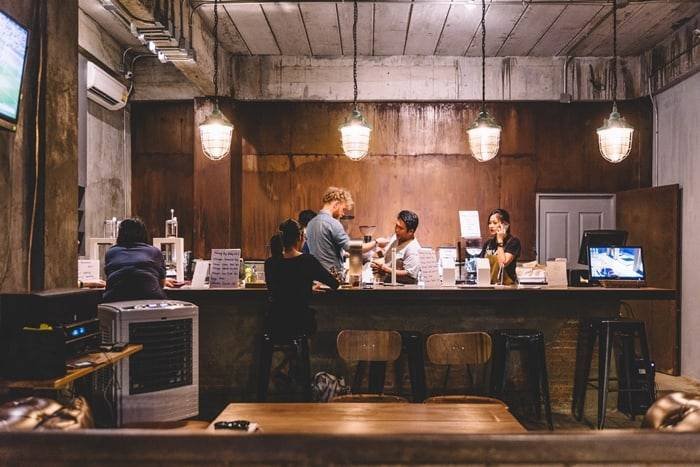 Qu&aacute;n c&agrave; ph&ecirc; với nhiều người ngồi v&agrave; đứng quanh quầy bar, &aacute;nh s&aacute;ng ấm &aacute;p v&agrave; kh&ocirc;ng gian nội thất gỗ.