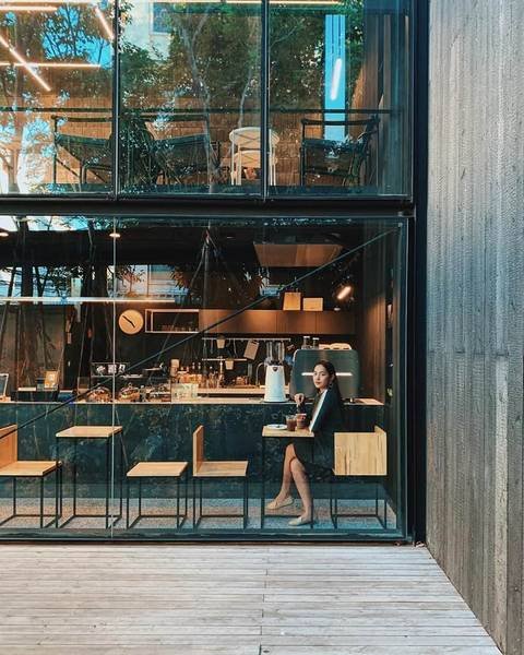 Người ngồi trong qu&aacute;n c&agrave; ph&ecirc; hiện đại với tường k&iacute;nh, xung quanh l&agrave; b&agrave;n ghế gỗ v&agrave; &aacute;nh s&aacute;ng tự nhi&ecirc;n.