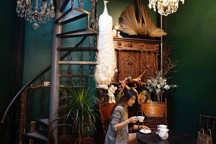 Người phụ nữ ngồi uống tr&agrave; trong kh&ocirc;ng gian trang tr&iacute; cổ điển với cầu thang xoắn v&agrave; c&acirc;y cối xung quanh.