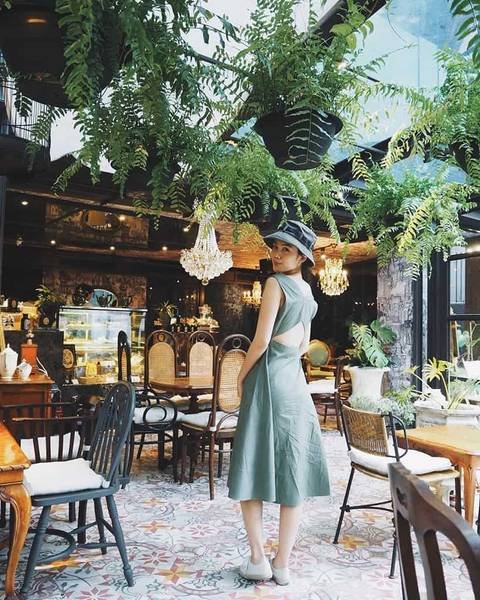 Một người mặc v&aacute;y xanh đứng trong qu&aacute;n c&agrave; ph&ecirc; trang tr&iacute; c&acirc;y xanh v&agrave; đ&egrave;n ch&ugrave;m, xung quanh c&oacute; nhiều b&agrave;n ghế.