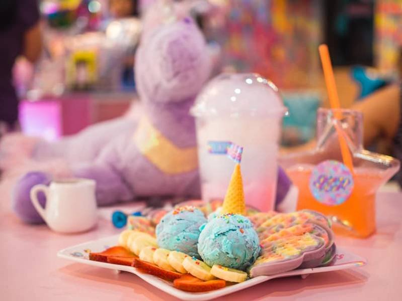 Đĩa b&aacute;nh quế v&agrave; kem m&agrave;u xanh, k&egrave;m l&aacute;t chuối v&agrave; d&acirc;u, b&ecirc;n cạnh l&agrave; đồ uống v&agrave; ch&uacute; ngựa b&ocirc;ng m&agrave;u t&iacute;m.