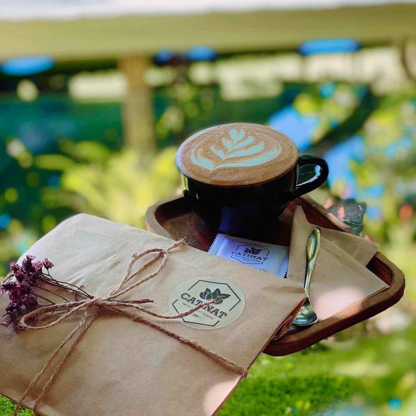 T&aacute;ch c&agrave; ph&ecirc; latte với tạo h&igrave;nh nghệ thuật tr&ecirc;n bọt, b&ecirc;n cạnh l&agrave; phong thư v&agrave; th&igrave;a bạc, tr&ecirc;n nền c&acirc;y xanh m&aacute;t.