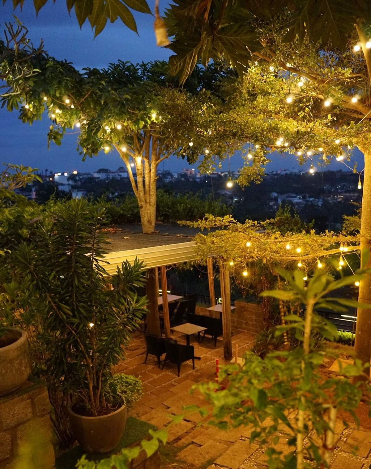 Khu vườn ngo&agrave;i trời với &aacute;nh đ&egrave;n v&agrave;ng lung linh, b&agrave;n ghế gỗ dưới t&aacute;n c&acirc;y, kh&ocirc;ng gian tho&aacute;ng đ&atilde;ng v&agrave;o buổi tối.
