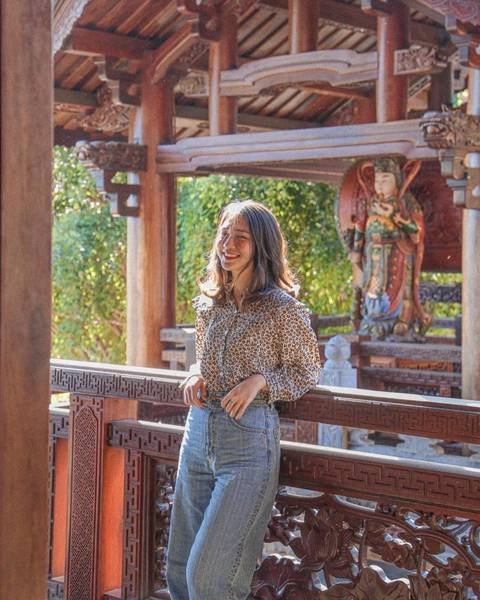 Người đang đứng tr&ecirc;n ban c&ocirc;ng gỗ với kiến tr&uacute;c truyền thống, ph&iacute;a sau l&agrave; tượng v&agrave; c&acirc;y cối xanh tươi.