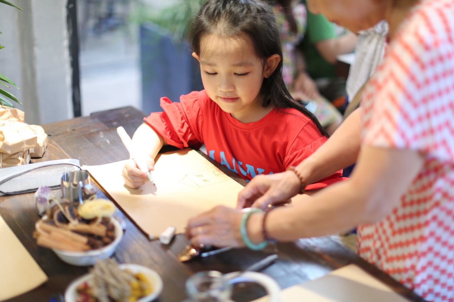 Một em nhỏ đang vẽ tr&ecirc;n giấy tại b&agrave;n, c&oacute; người lớn hướng dẫn b&ecirc;n cạnh. Tr&ecirc;n b&agrave;n c&oacute; nhiều vật dụng kh&aacute;c nhau.