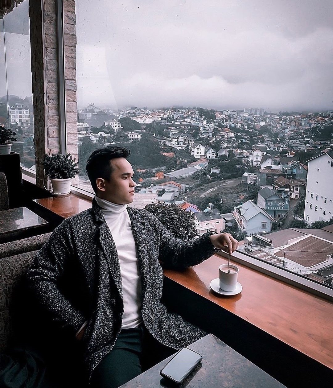 Người ngồi uống c&agrave; ph&ecirc; b&ecirc;n cửa sổ lớn, nh&igrave;n ra cảnh th&agrave;nh phố m&ugrave; sương b&ecirc;n ngo&agrave;i.