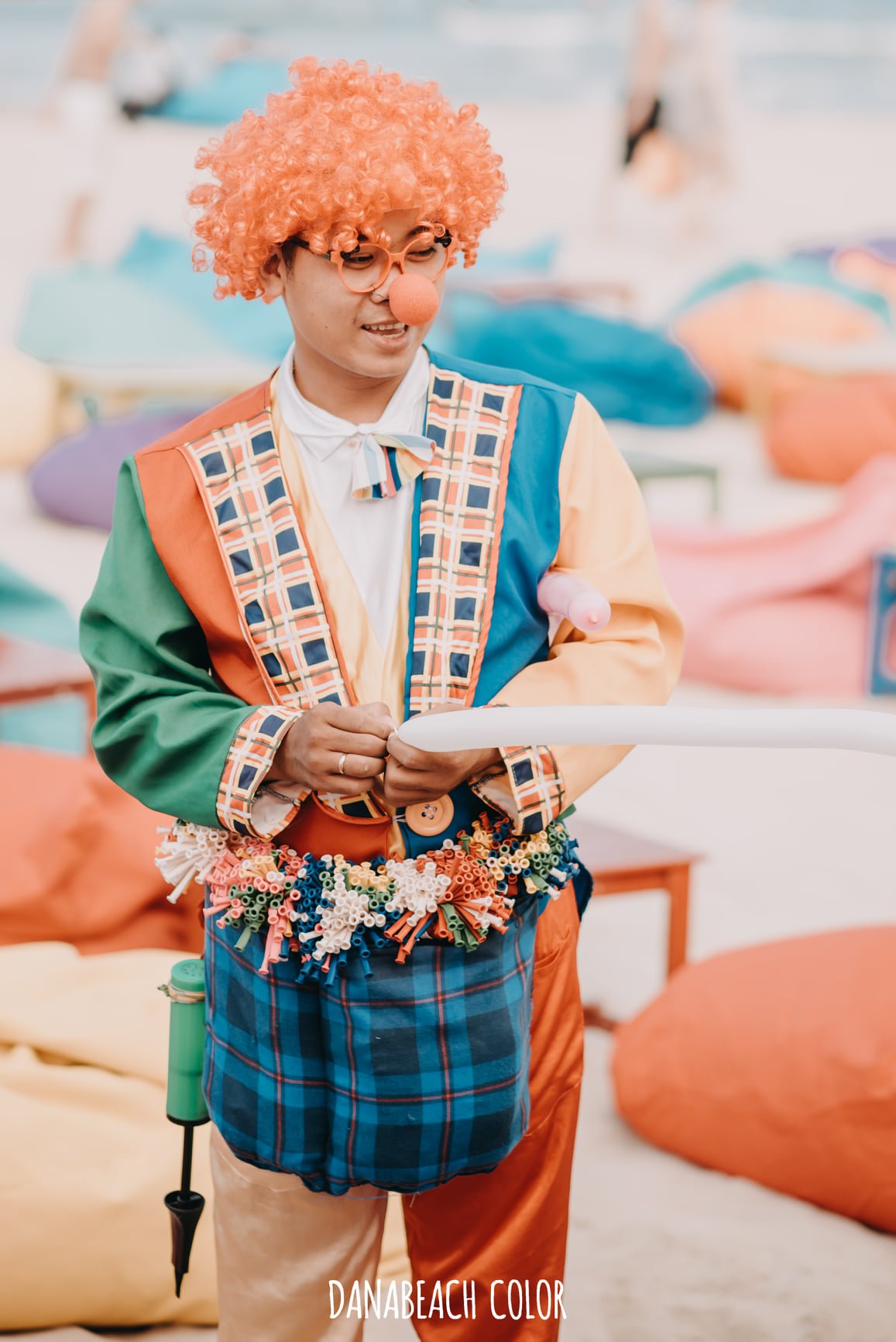 Người mặc trang phục ch&uacute; hề đầy m&agrave;u sắc, đứng tr&ecirc;n b&atilde;i biển với những t&uacute;i đầy ống h&uacute;t v&agrave; b&oacute;ng bay.