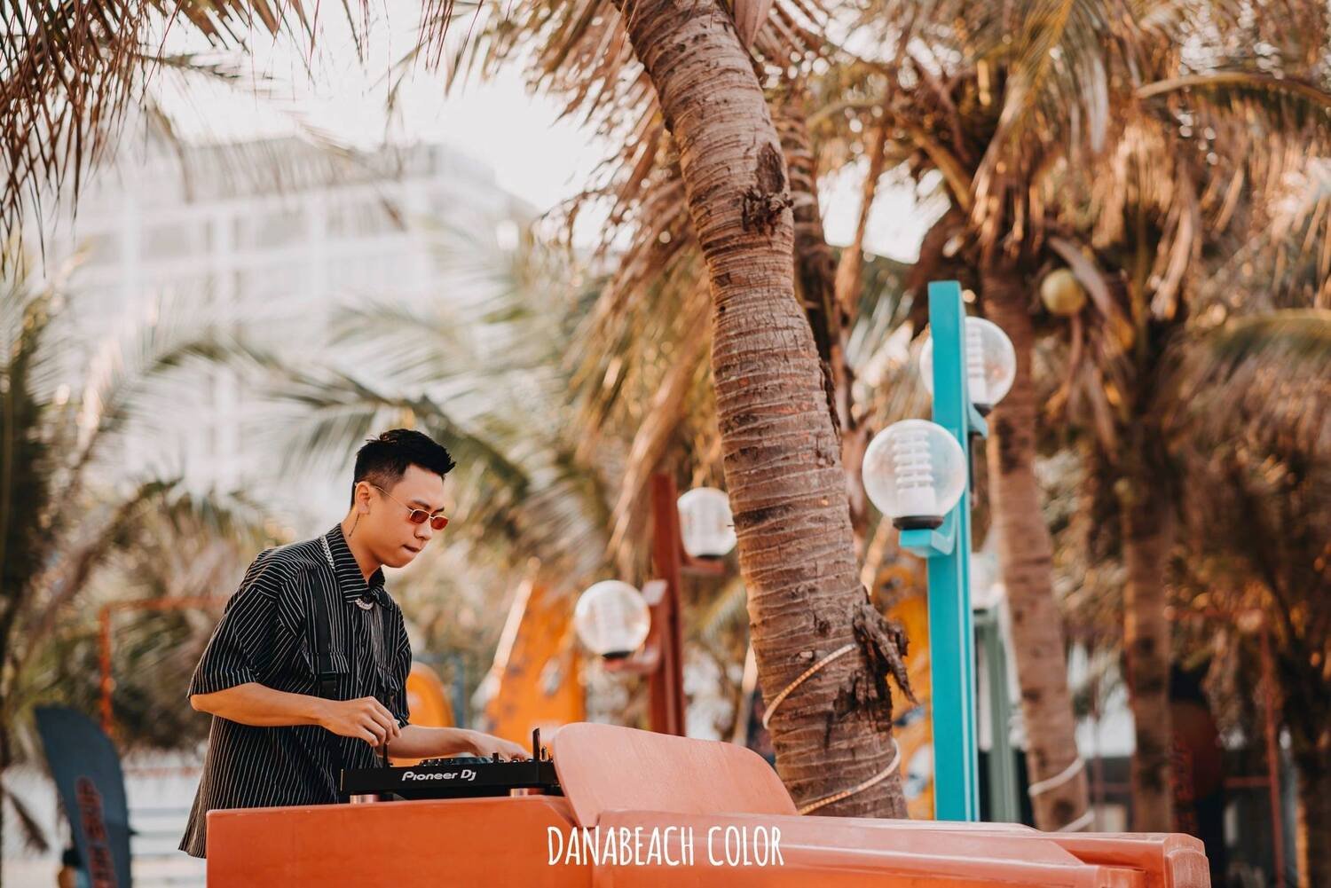 Người đ&agrave;n &ocirc;ng điều chỉnh b&agrave;n DJ ngo&agrave;i trời, xung quanh l&agrave; c&acirc;y cọ v&agrave; đ&egrave;n trang tr&iacute;.