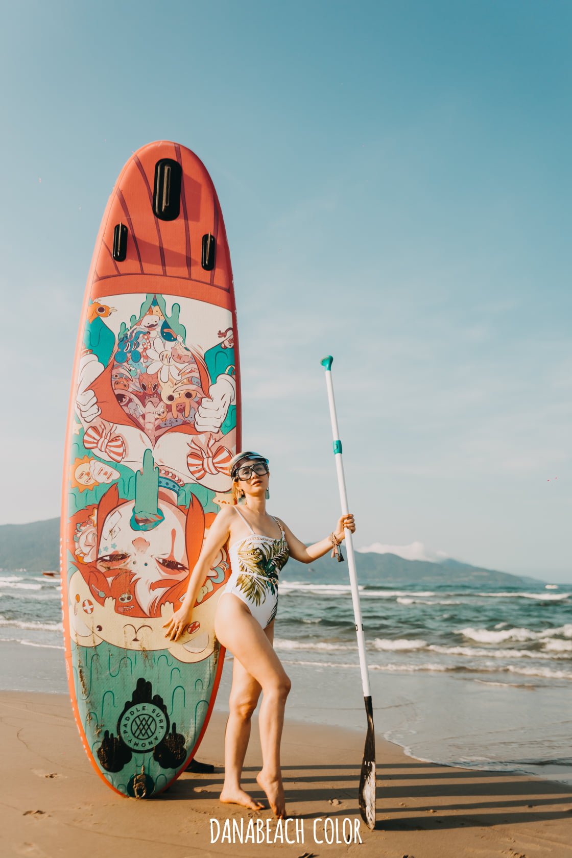 Người đứng tr&ecirc;n b&atilde;i biển, cầm m&aacute;i ch&egrave;o, b&ecirc;n cạnh v&aacute;n lướt s&oacute;ng c&oacute; họa tiết nhiều m&agrave;u sắc.