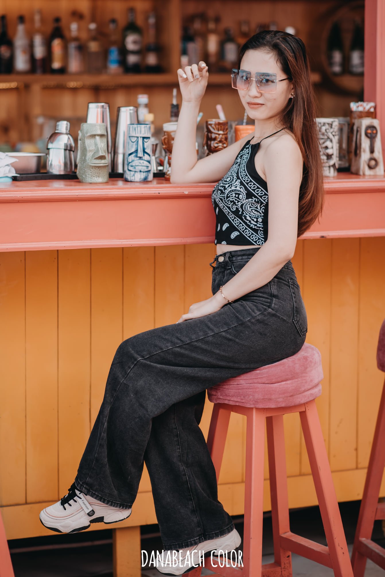 Người phụ nữ ngồi tr&ecirc;n ghế cao tại quầy bar ngo&agrave;i trời, đeo k&iacute;nh m&aacute;t v&agrave; mặc trang phục thời trang.