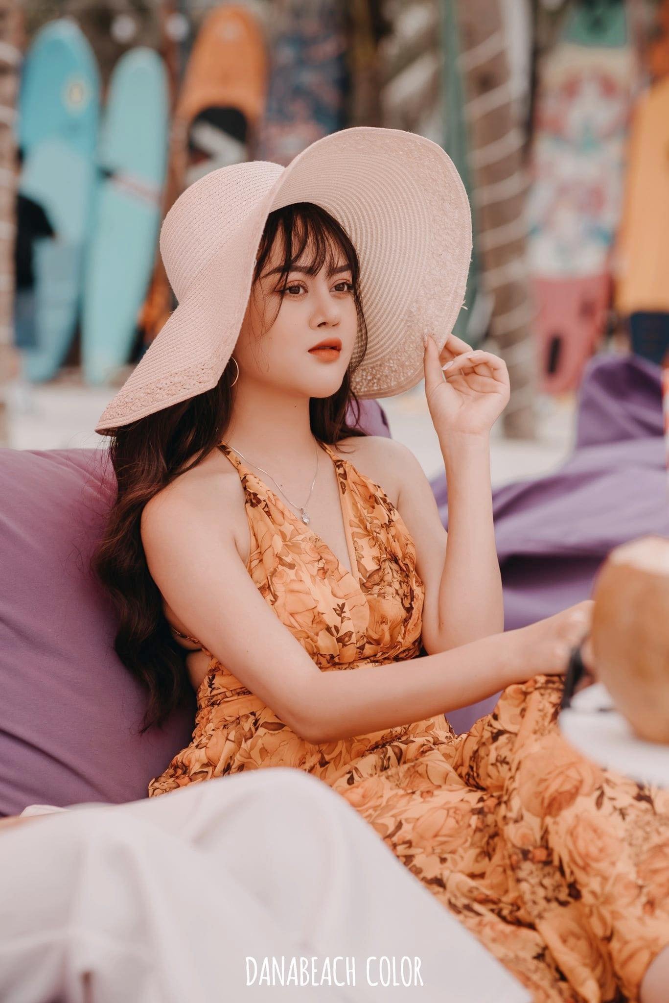 Người phụ nữ đội n&oacute;n rộng v&agrave;nh v&agrave; mặc v&aacute;y hoa, ngồi thư gi&atilde;n tr&ecirc;n ghế b&atilde;i biển với ph&ocirc;ng nền v&aacute;n lướt s&oacute;ng.