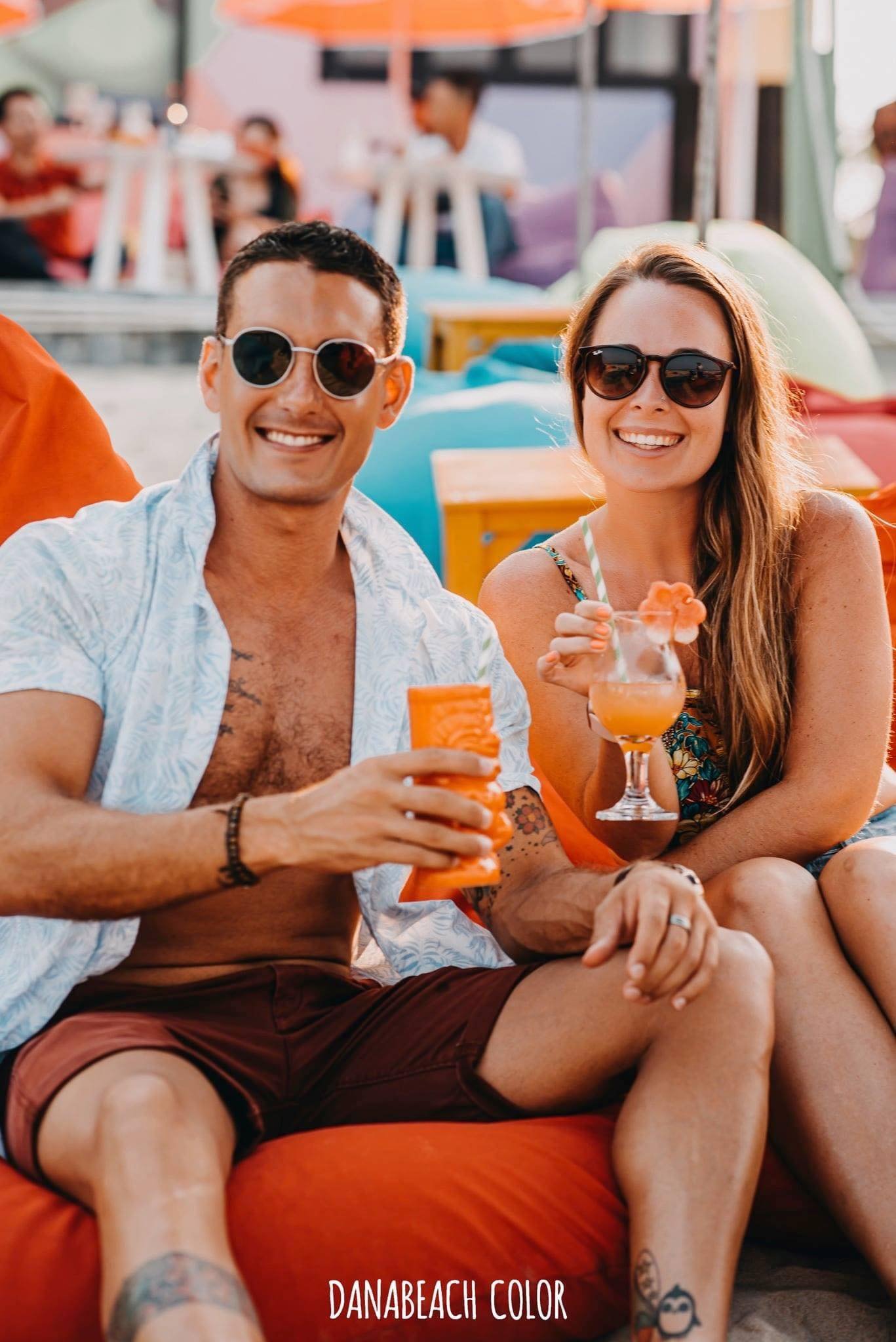 Hai người ngồi tr&ecirc;n ghế lười, cười tươi v&agrave; cầm ly nước, trong bối cảnh b&atilde;i biển s&ocirc;i động.